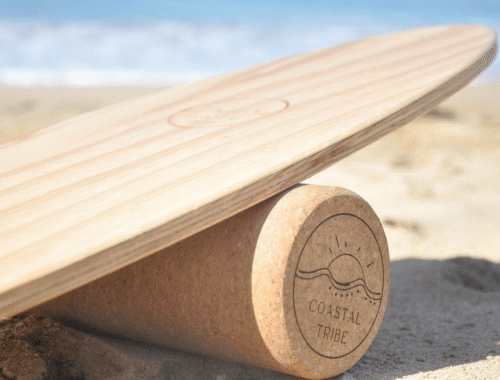 Coastal Tribe surf balance board "The Shredder", close-up image of the beautifully crafted timber board resting on the cork roller on a sandy beach with the ocean in the background