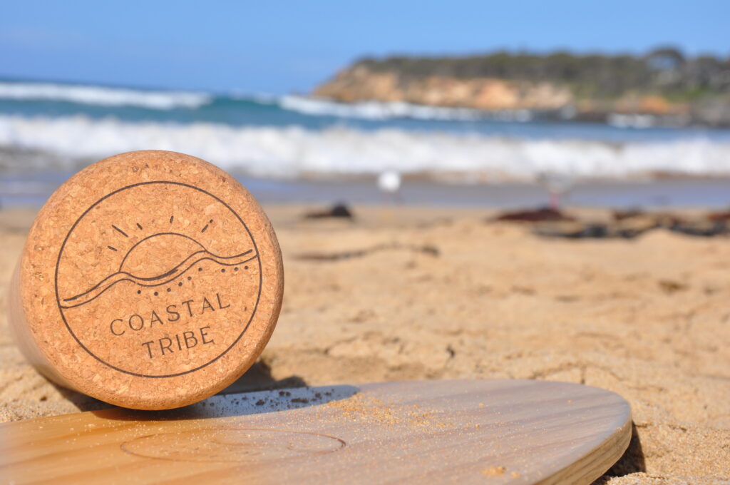 Coastal Tribe Surf Balance Board, The Shredder, on a beach with sand sprinkled across the timber board, ocean in the background, looking stunning.